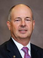 Middle-aged man with short hair in a suit and red tie, standing against a plain brown background.